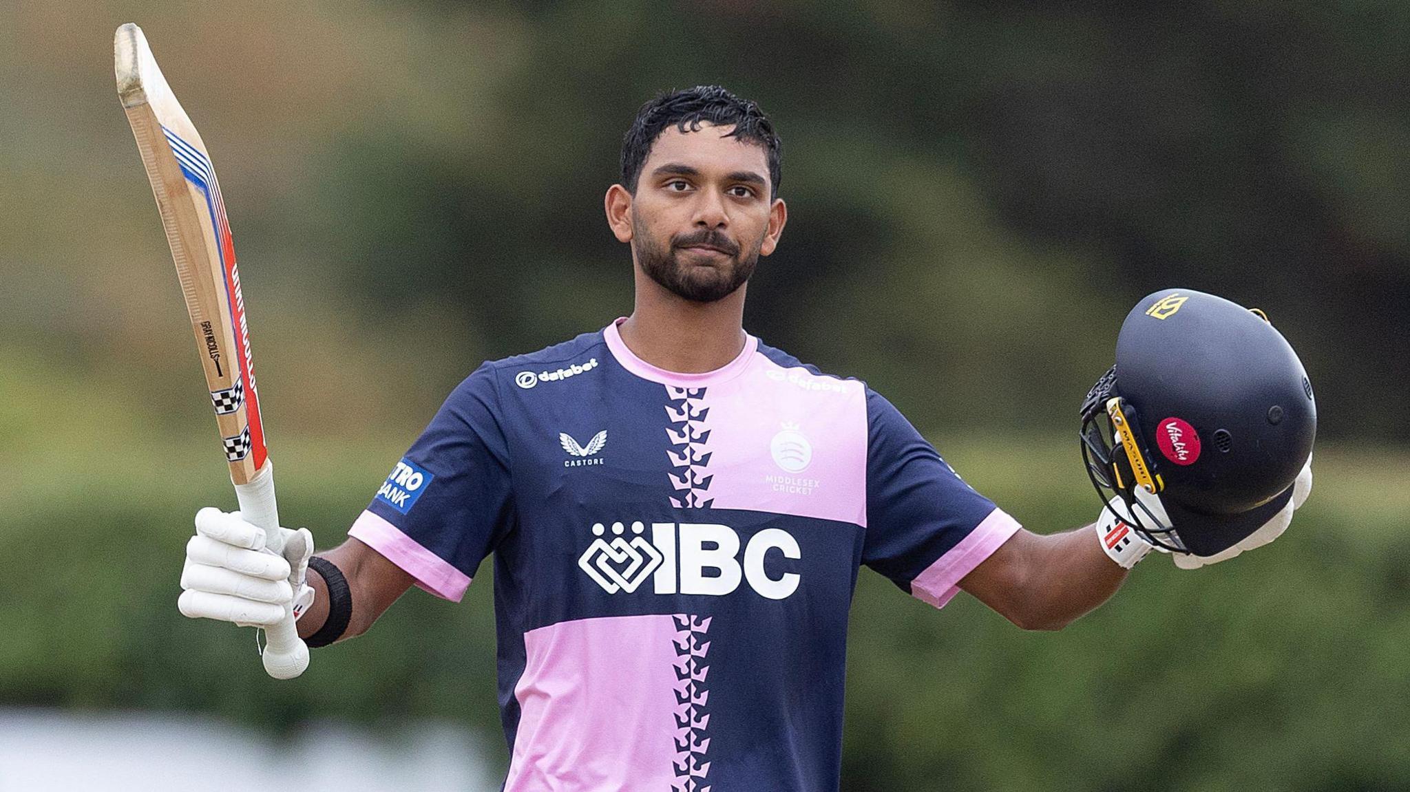 Nathan Fernandes holds up his bat and helmet as he celebrates scoring a century for Middlesex. He is wearing a pink and navy blue quartered shirt.