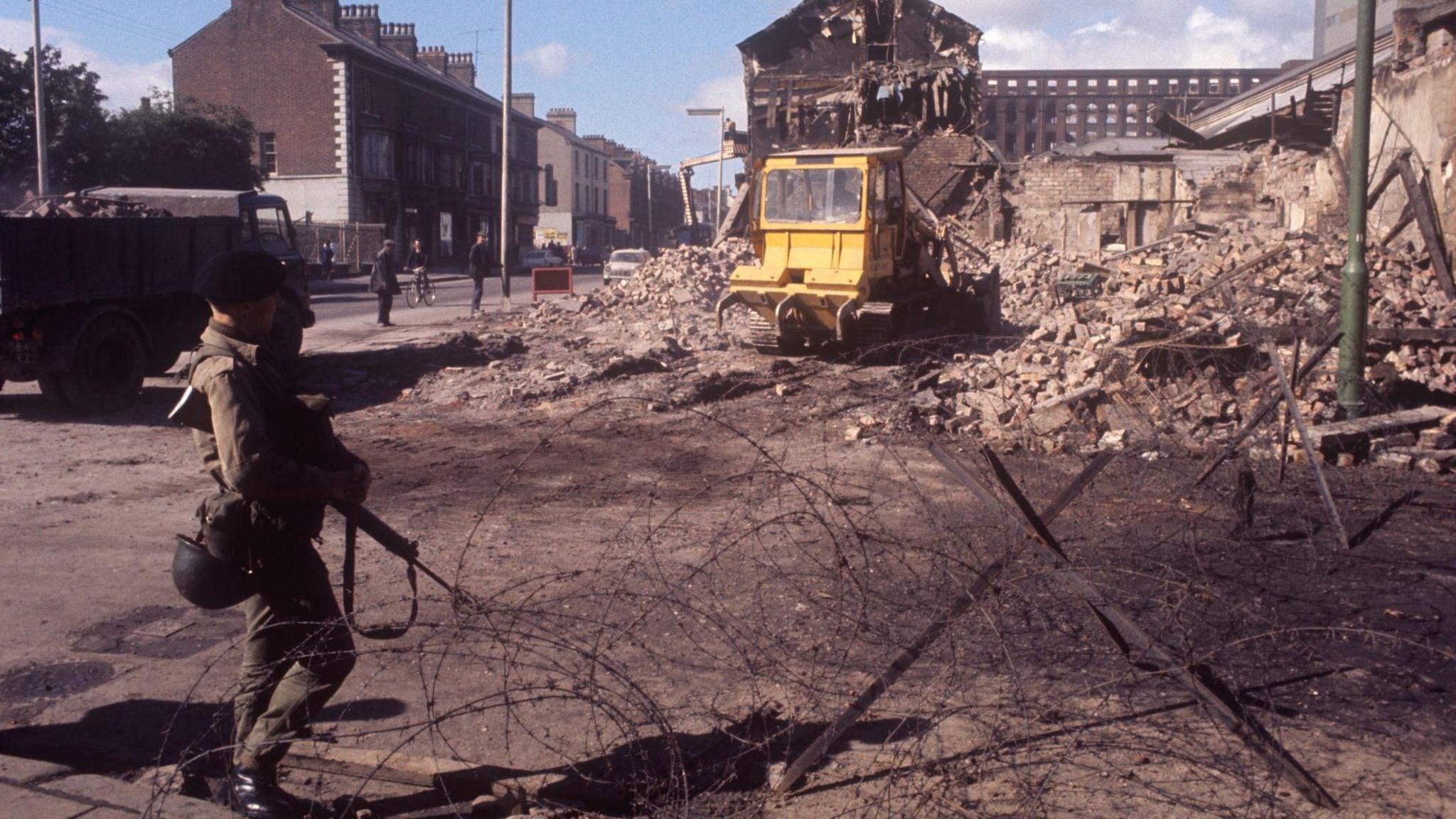 A British soldier stands at barbed wire. In front of him is a pile of rubble and a yellow forklift truck. It's a picture from the Troubles in NI.