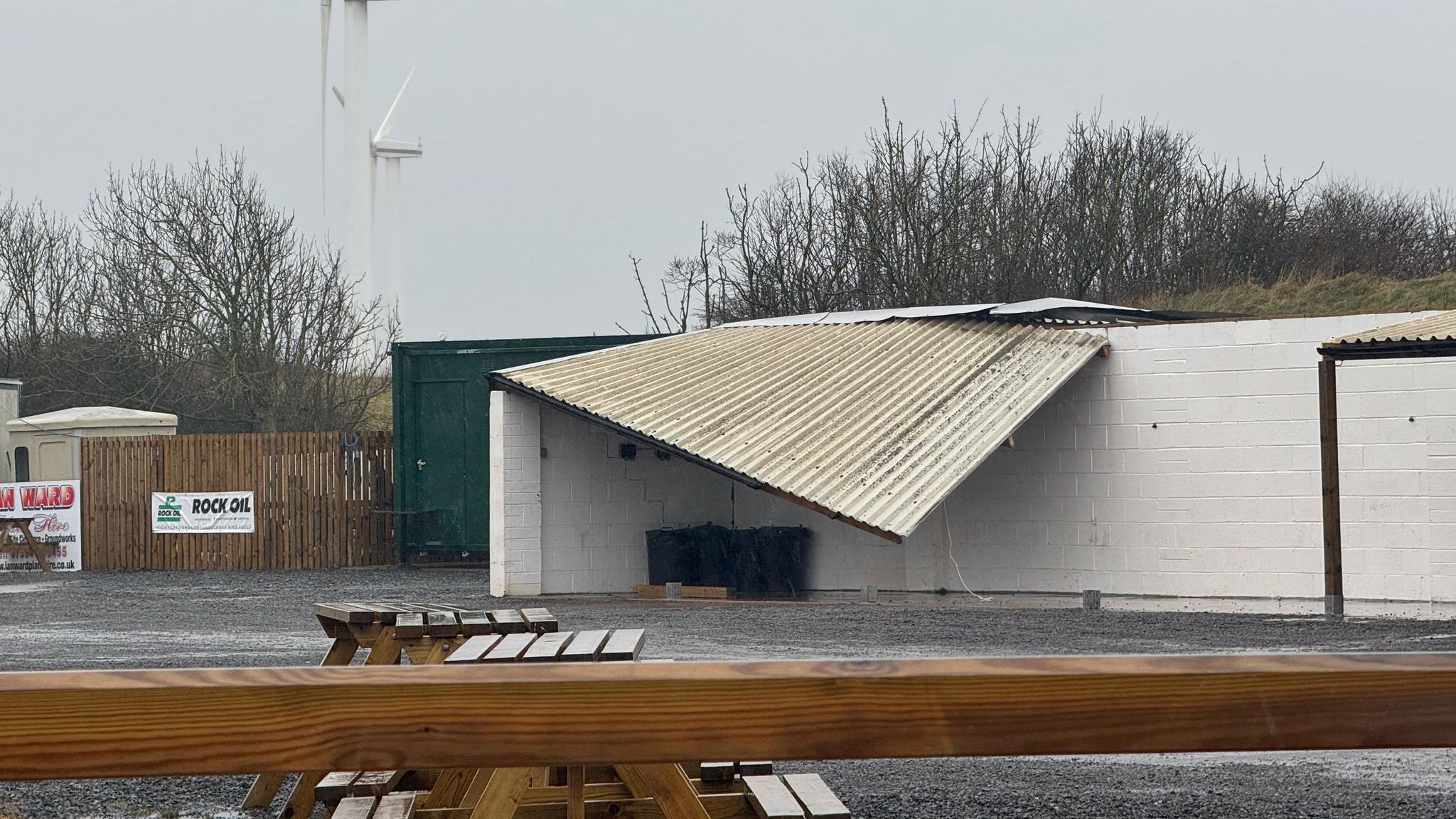 A single-storey white brick building with no windows. Its corrugated has been ripped off and is hanging over the edge of the building.