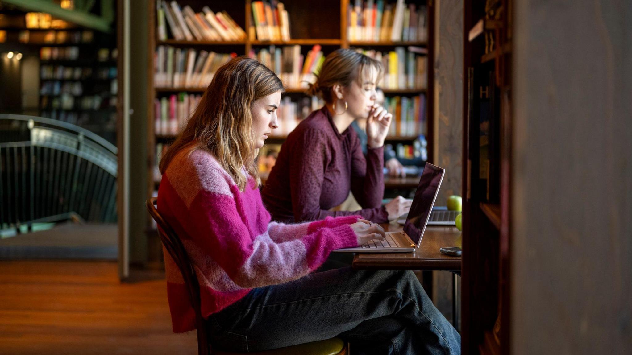 Two young women sit at laptops with library shelves full of books behind them