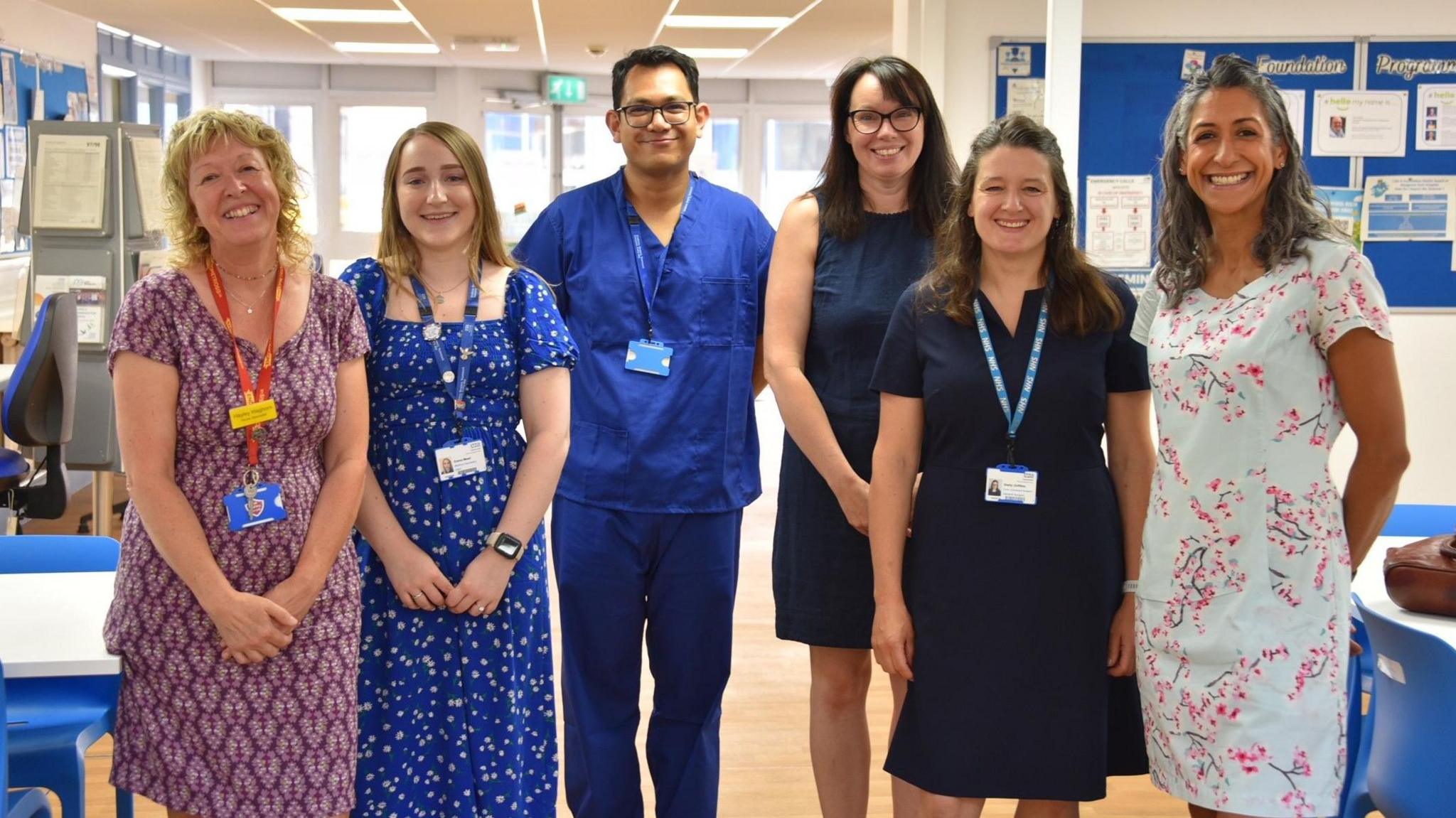 A group of NHS staff made up of five women and one man smile as they stand in a line facing the camera. Most of them are wearing casual clothes, although the man is in blue medical scrubs. They are in a hospital ward with a light-coloured floor
