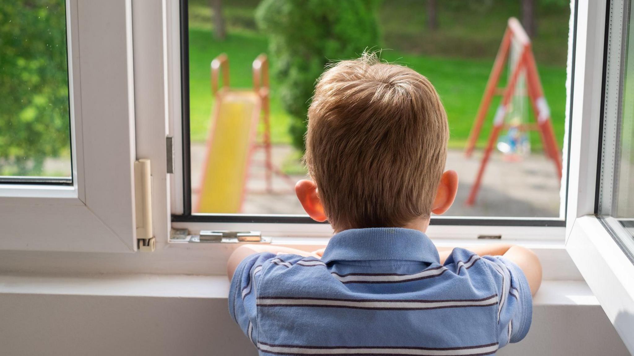 a little boy with his back to the camera, with arms folded on a window ledge looking out at a garden with a children's slide and swing