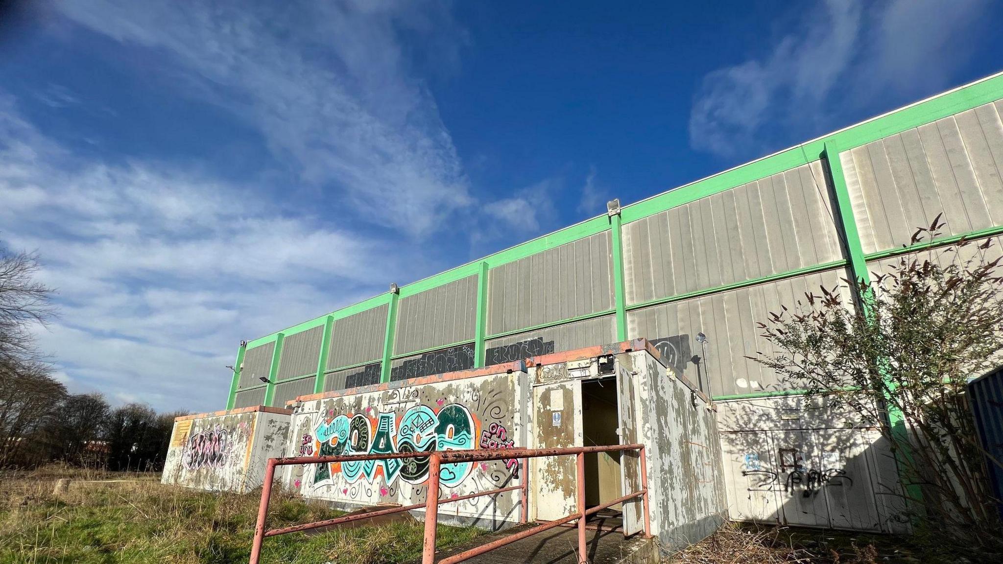 The front of a warehouse-type building with large green metal bars. A ramp leads up to an open door, with the porch area covered in graffiti. There is overgrown grass round the building