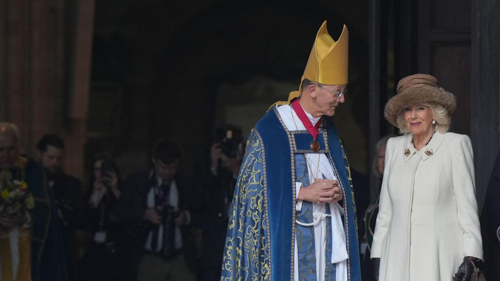 Bishop Inge, wearing a gold mitre and a blue robe, stands looking at The Queen who is wearing a cream-coloured coat and a brown hat.