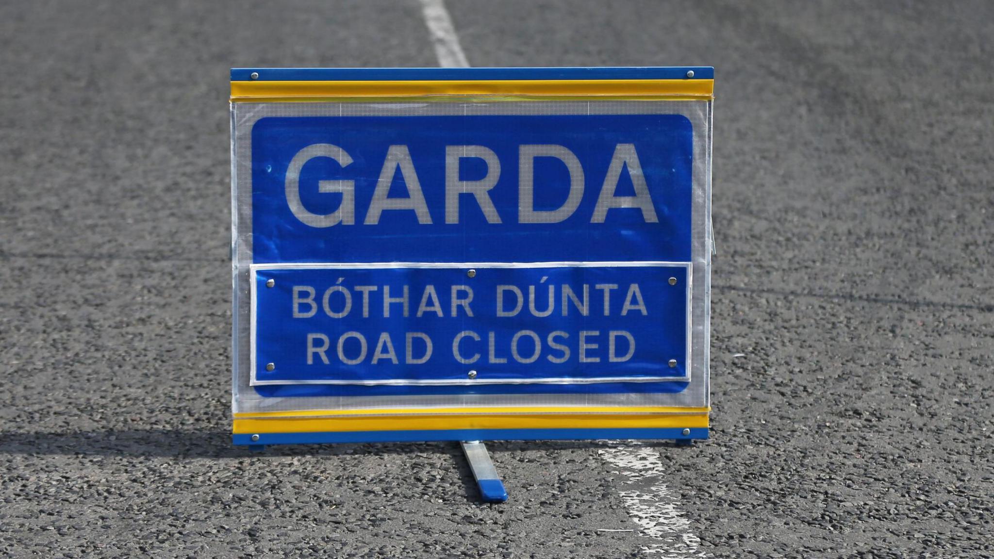 A blue bilingual road sign which reads:" Garda - bothar dunta - road closed."
The sign in sited in the middle of an empty road, under a cordon made of blue and white tape