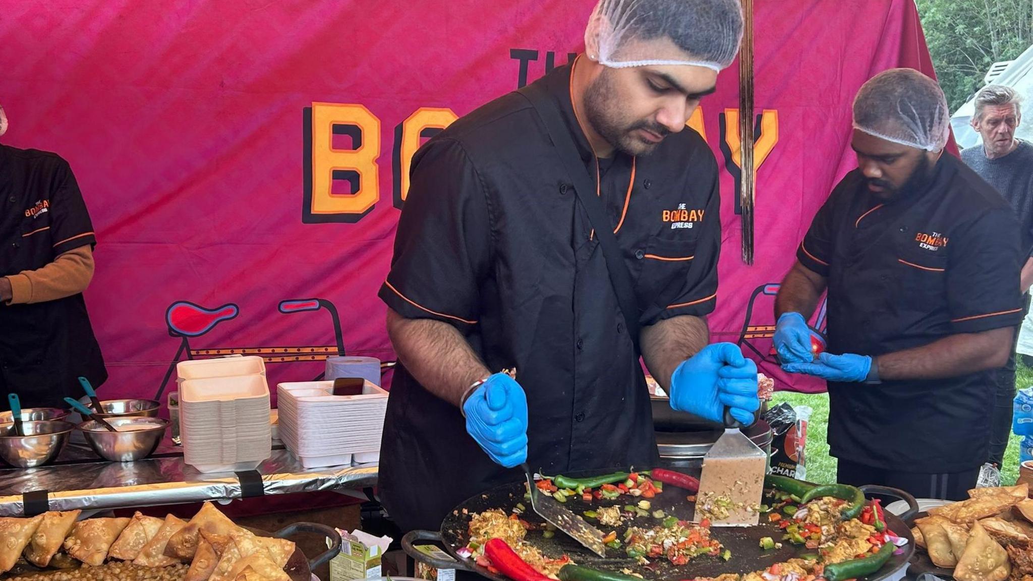 Two men wearing hairnets and wearing blue plastic gloves frying chillies while preparing Indian street food. There is a purple background and food cartons and dishes behind them. 