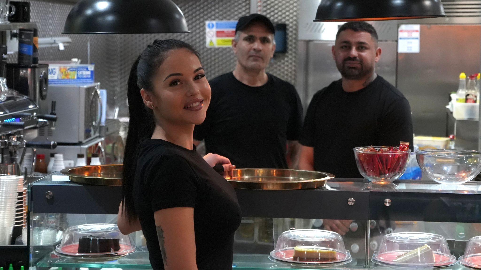 Brigitta Lantos is looking backwards and smiling at the camera. She has long, dark hair tied back and is wearing a black T-shirt. Two men also in black tops are standing on the other side of a serving counter in a restaurant