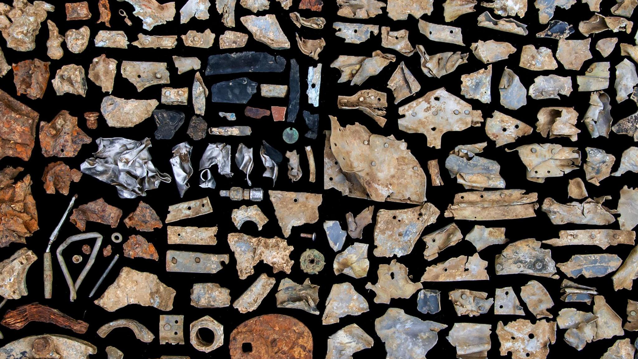 Fragments of material found at a plane crash site are lined up on a black background. There are various bits of metal and bolts