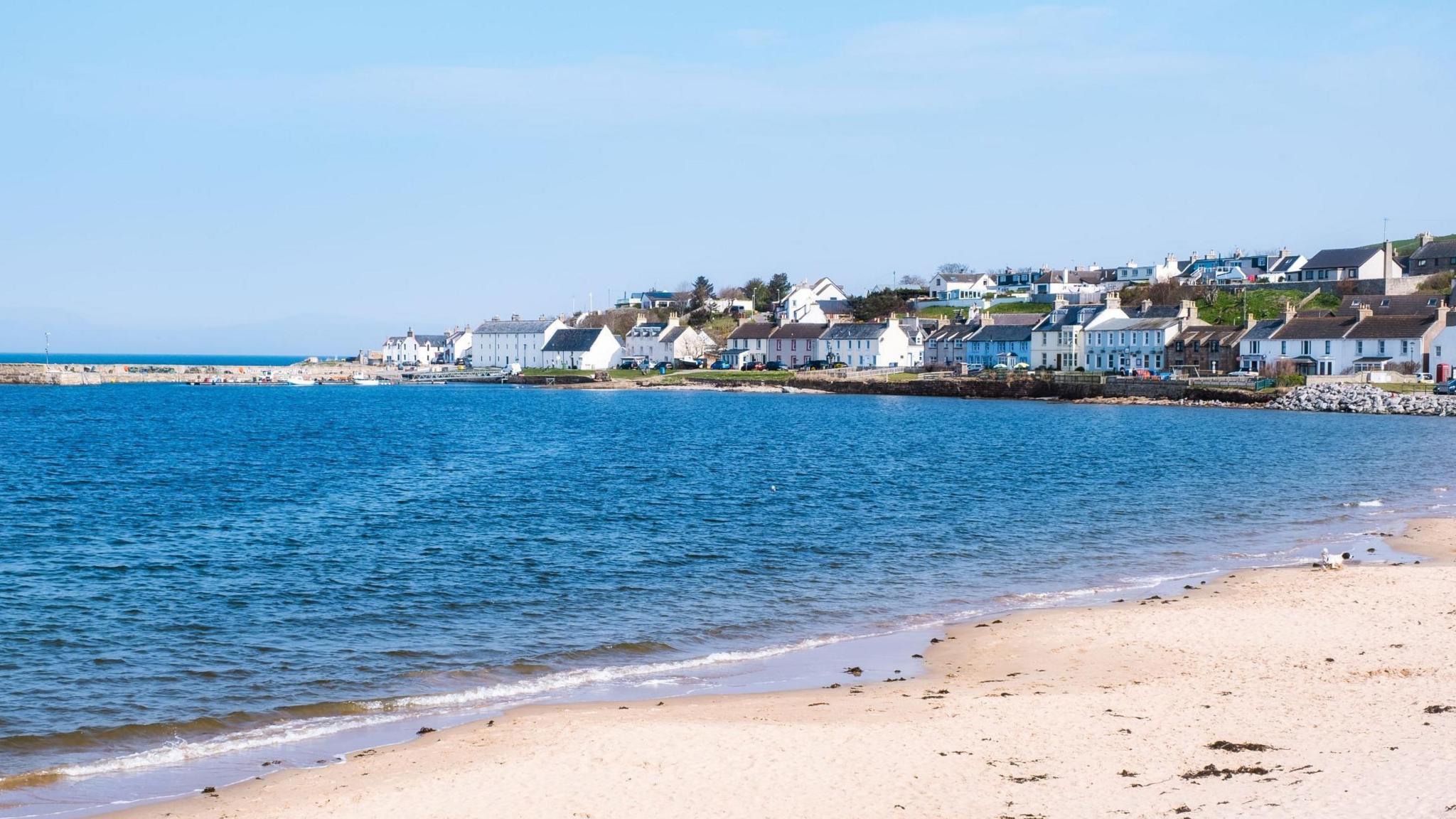 Portmahomack is a coastal village with white-walled houses and buildings related to the fishing industry and its harbour. In the foreground is a sandy beach. The picture was taken on a sunny day and the sky is blue and cloudless.