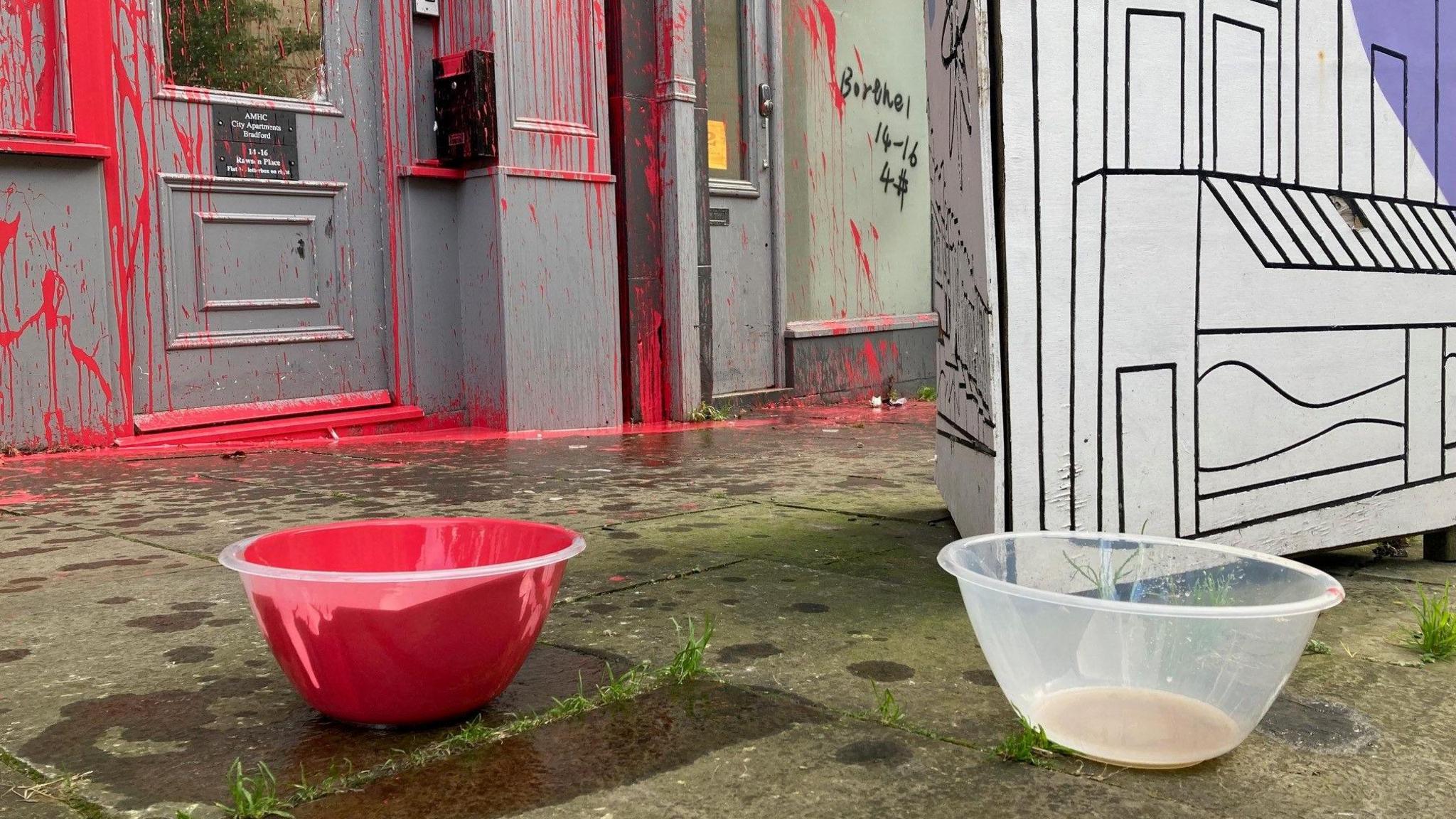 A plastic bowl with red paint in it on a pavement with a similar bowl containing a light brown liquid next to it. In the background is a doorway covered in red paint. 