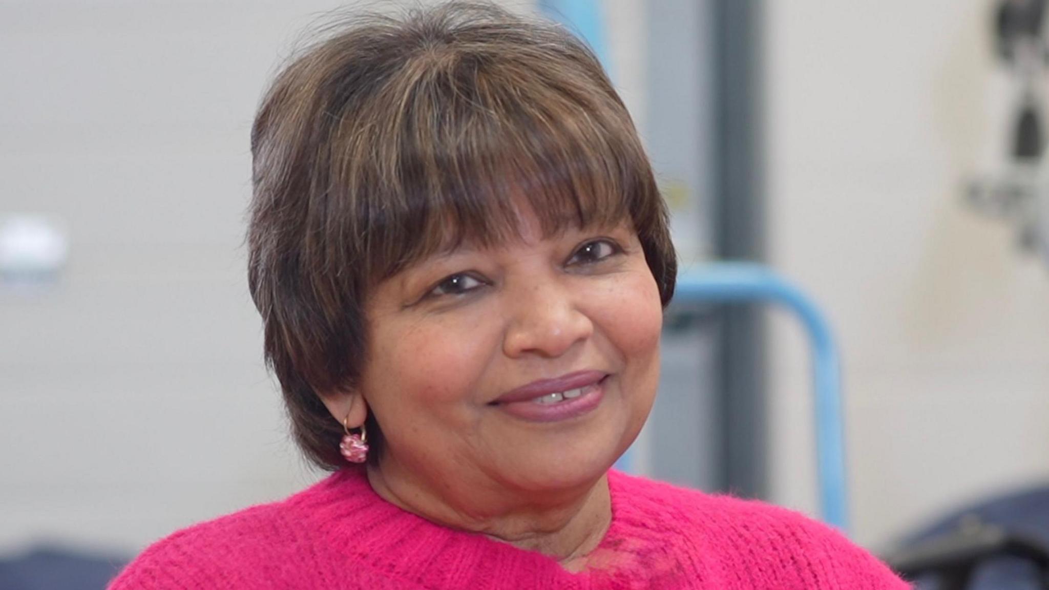 Prema Fairburn-Dorai smiles at the camera. She has a brown hair that is cut into a pixie bob with a fringe. She is wearing a pink jumper and has pink earrings in.