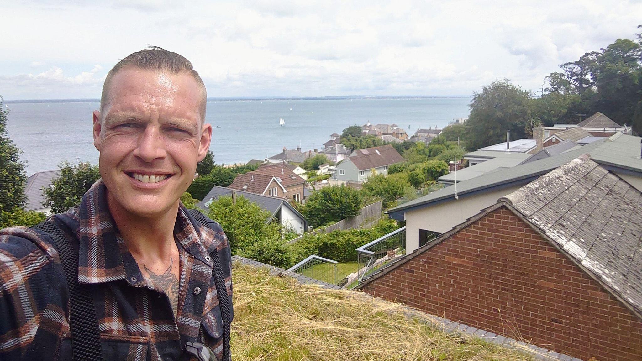 Tim Parry takes a selfie on a hilltop overlooking the coast off the Isle of Wight. He has short, light brown hair and wears a tartan purple and red shirt.