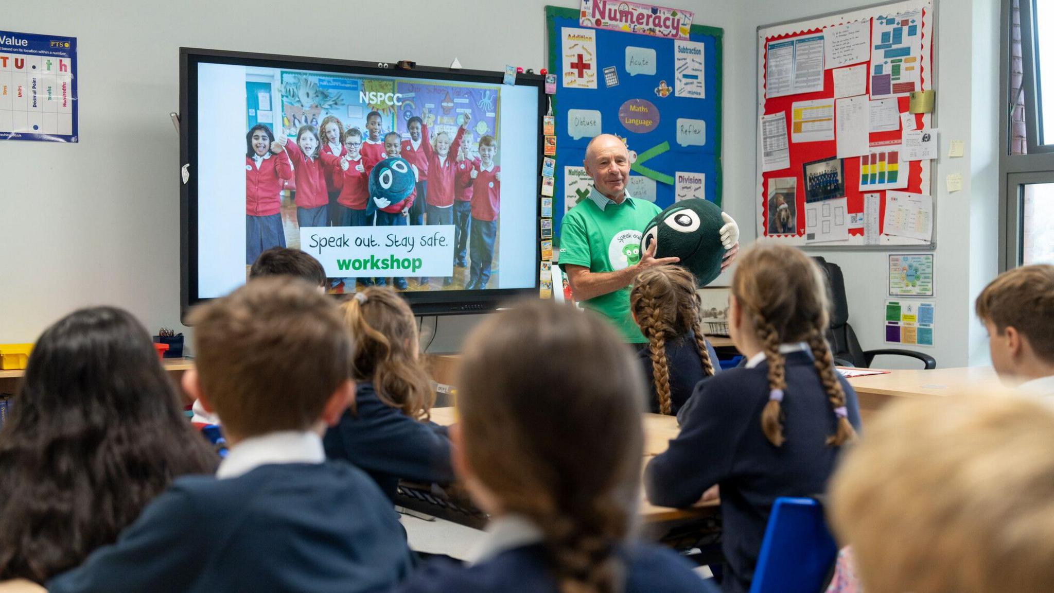 An NSPCC volunteer holds a mascot while talking to a class of primary school pupils during a Speak Out Stay Safe workshop. A presentation is up on the interactive whiteboard. The volunteer is wearing a green t-shirt.