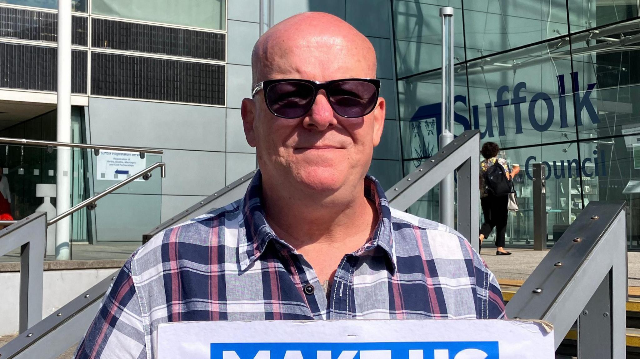 Mark Jones stands outside Suffolk County Council's headquarters in a large glass building. He is bald and wears sunglasses along with a navy, white and red chequered shirt.