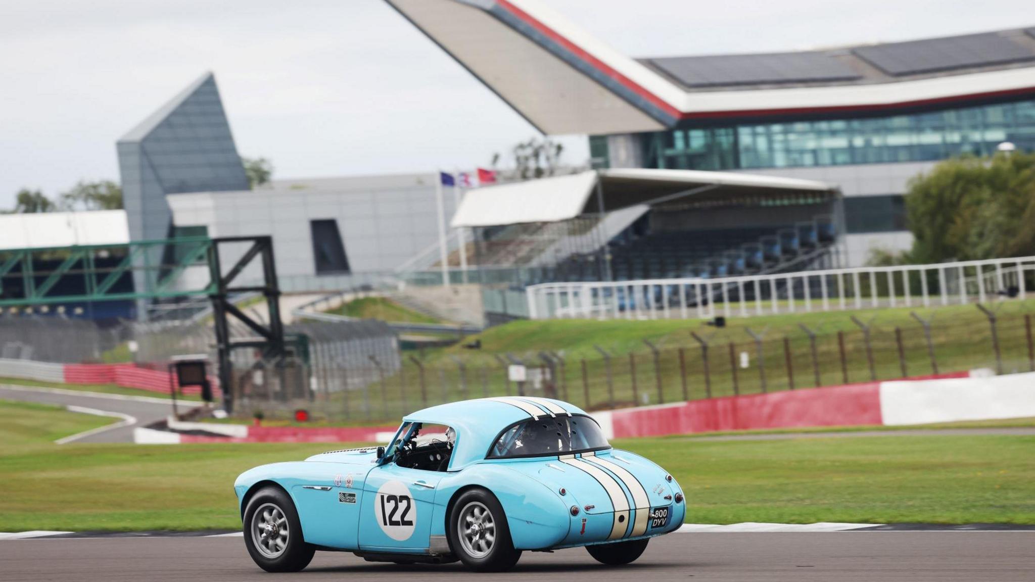 A blue 1960s sports car on the track. Behind it is a grandstand and building around the circuit.