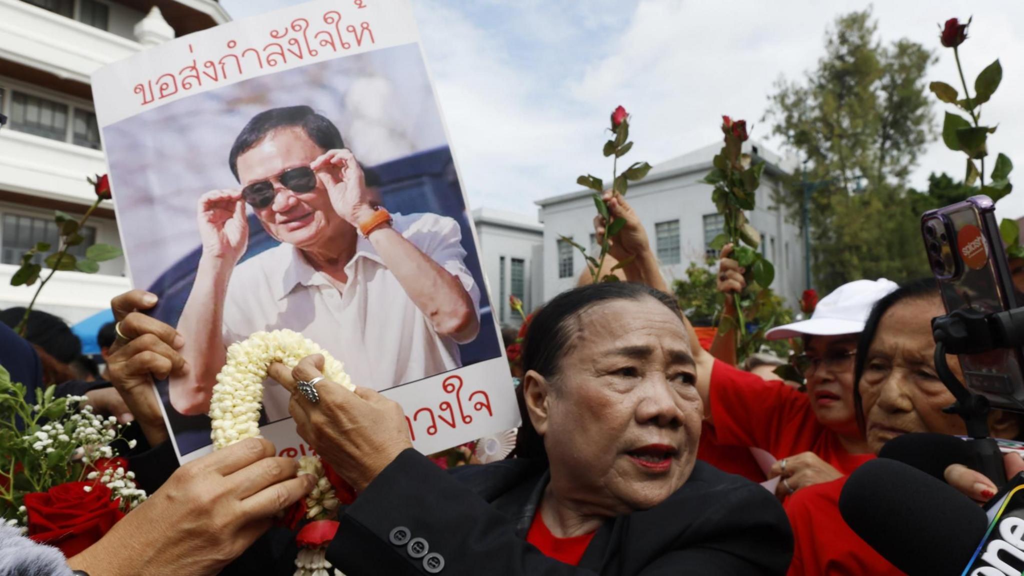 A woman wearing a black jacket and red shirt, surrounded by others wearing red shirts, holds up a poster of Thaksin depicting him wearing sunglasses and a white shirt
