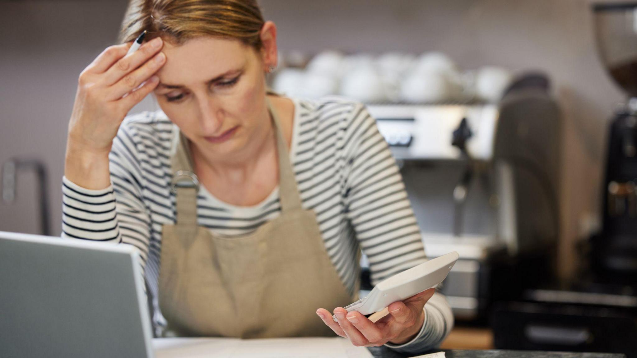 Worried Female Owner Of Coffee Shop Looking Through Bills Using Laptop And Calculator