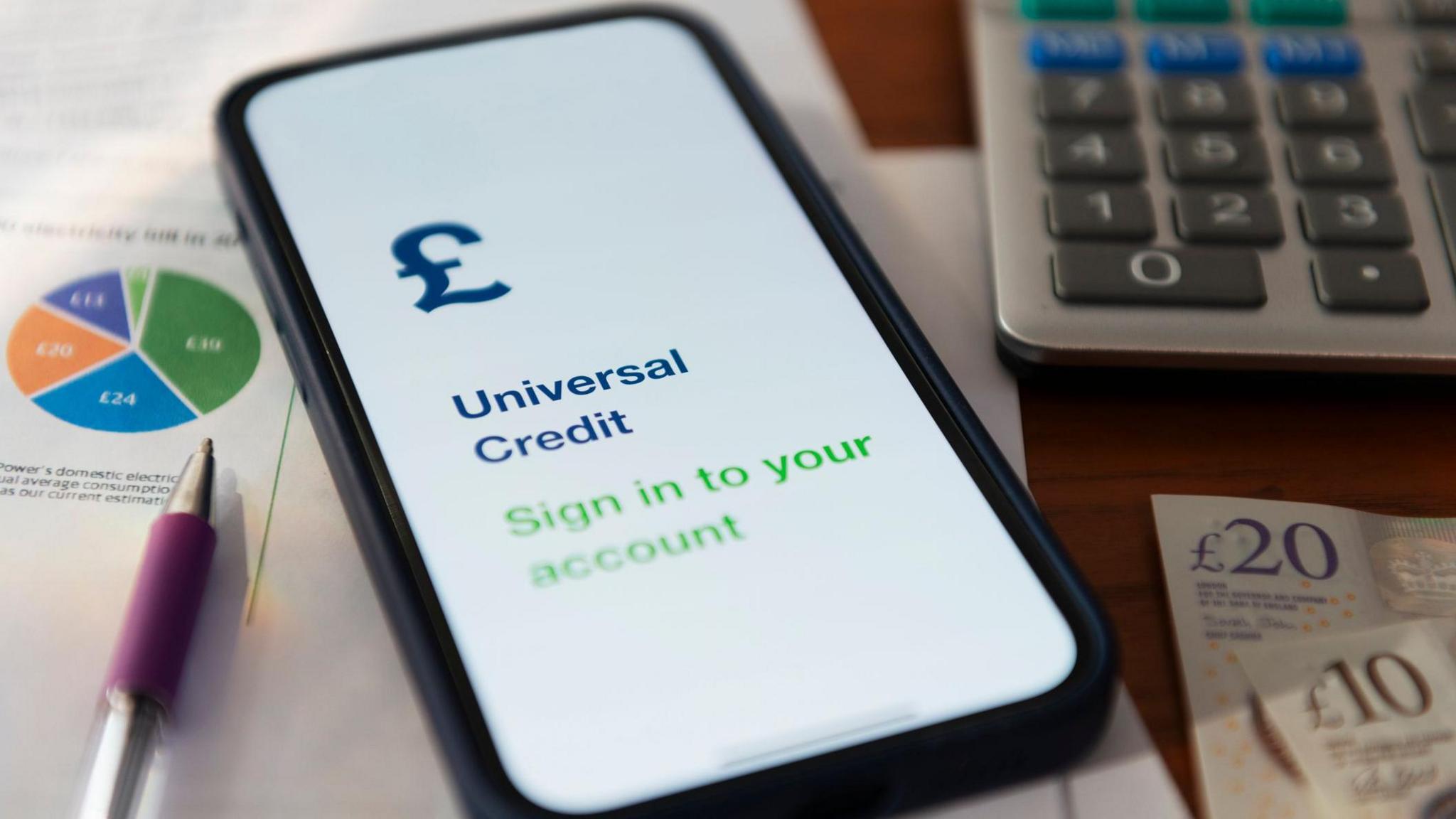 A shot of several objects including a pen, a smartphone open on a page to log in to a universal credit account, a calculator and two cash notes of £20 and £10 respectively.