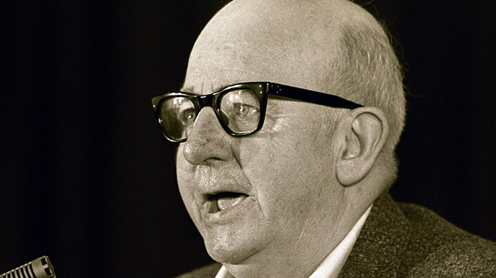 A black and white image of Joe Cahill. He is bald and wearing glasses speaking in front of a microphone.