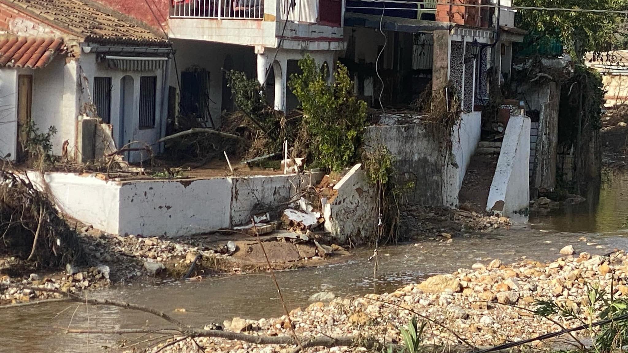 Water flooding past damaged houses on a Spanish mountain. Parts of the garden walls have collapsed and there is debris and foliage around.