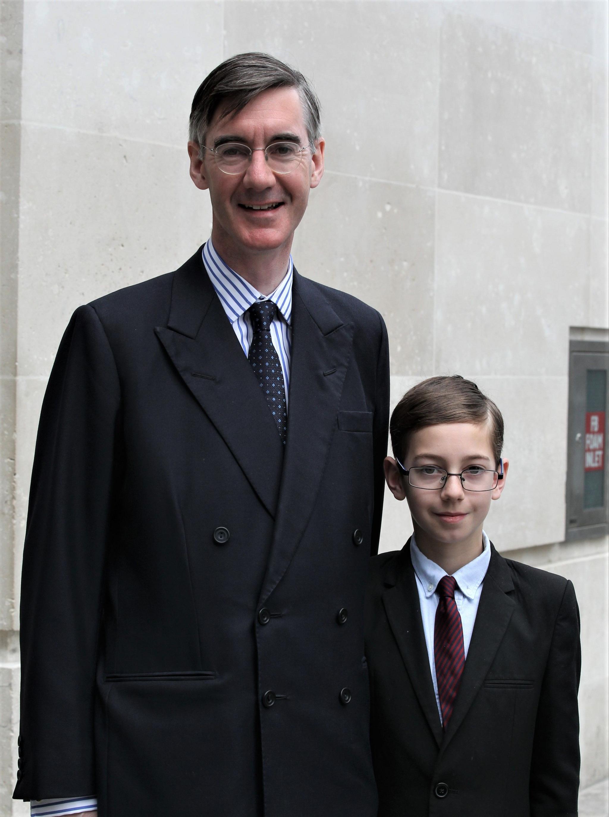 Jacob Rees-Mogg with his eldest son, Peter Theodore Alphege in 2018. They are both wearing black suits, shirts and ties. Both also wear glasses.