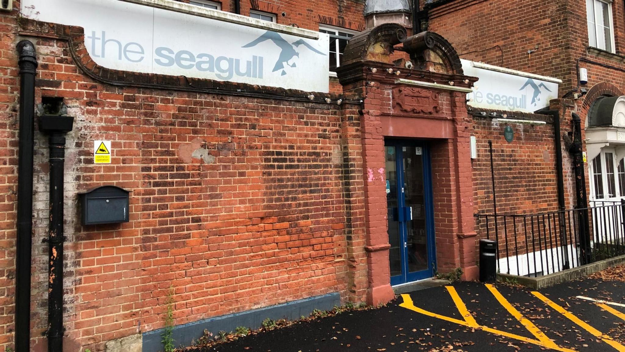 A view of Seagull Theatre in Pakefield, Lowestoft. An orange brick building with two large white signs either side of a blue doorway entrance that say "The Seagull".
