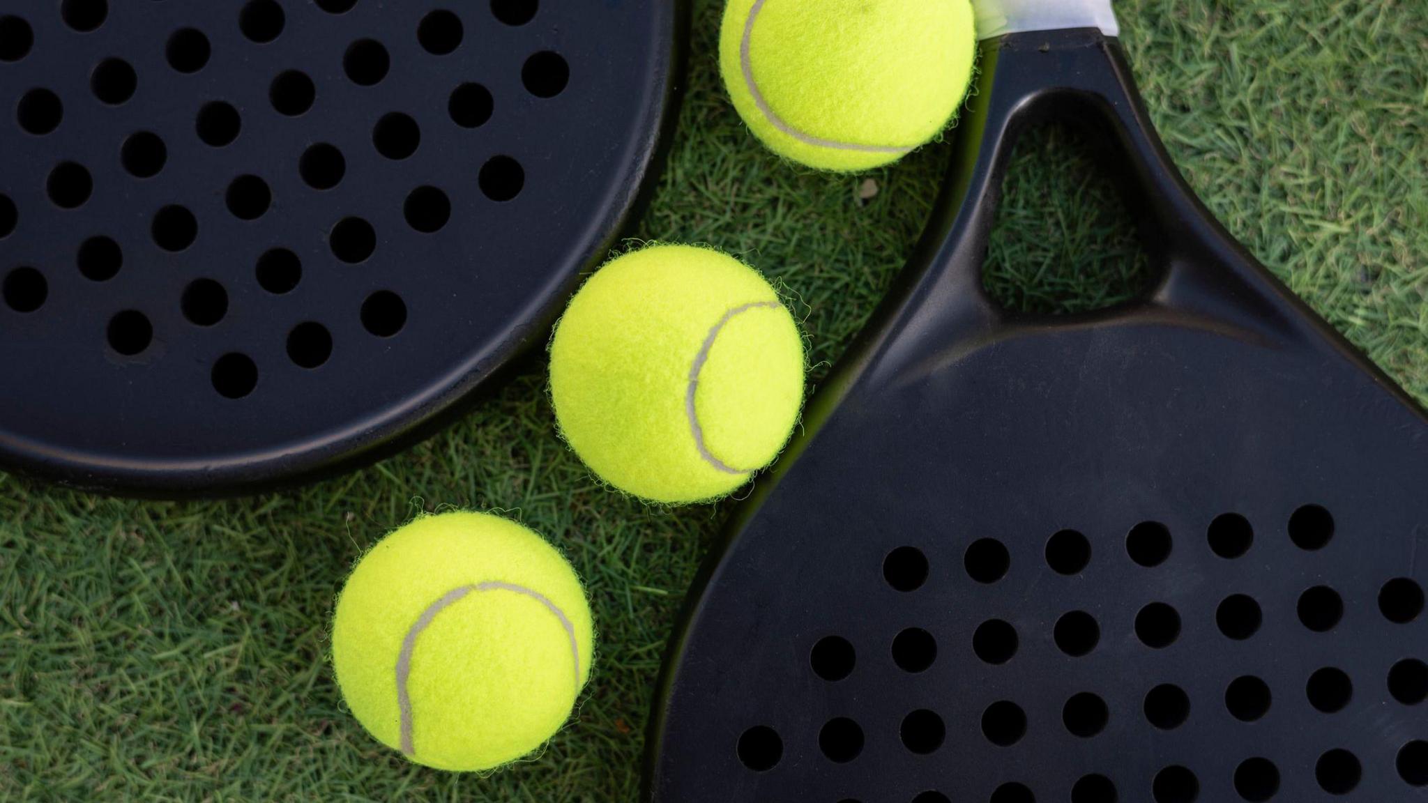 Three tennis balls and two padel rackets pictured on grass. 