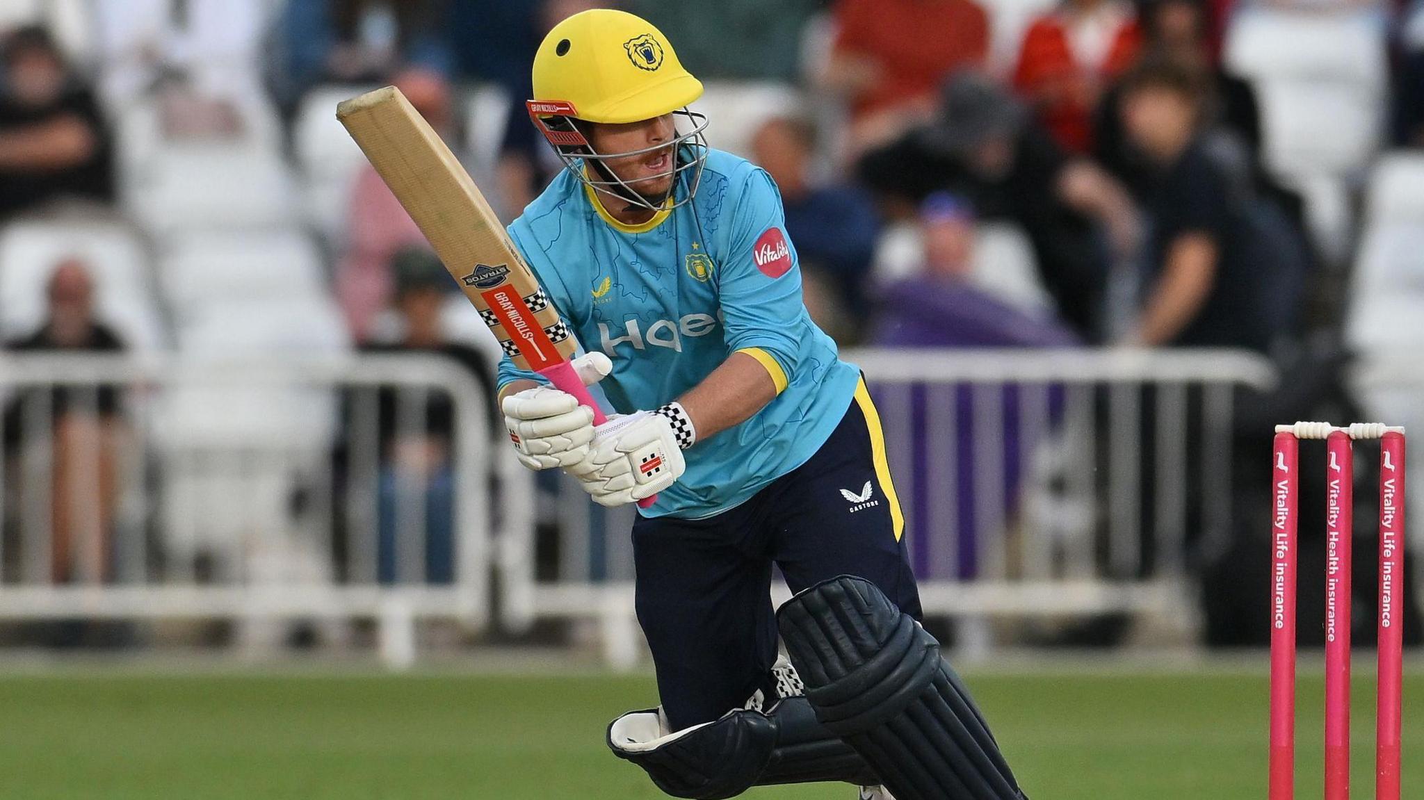 Warwickshire batter Alex Davies, wearing a blue shirt and a yellow helmet, holds his bat after playing a shot
