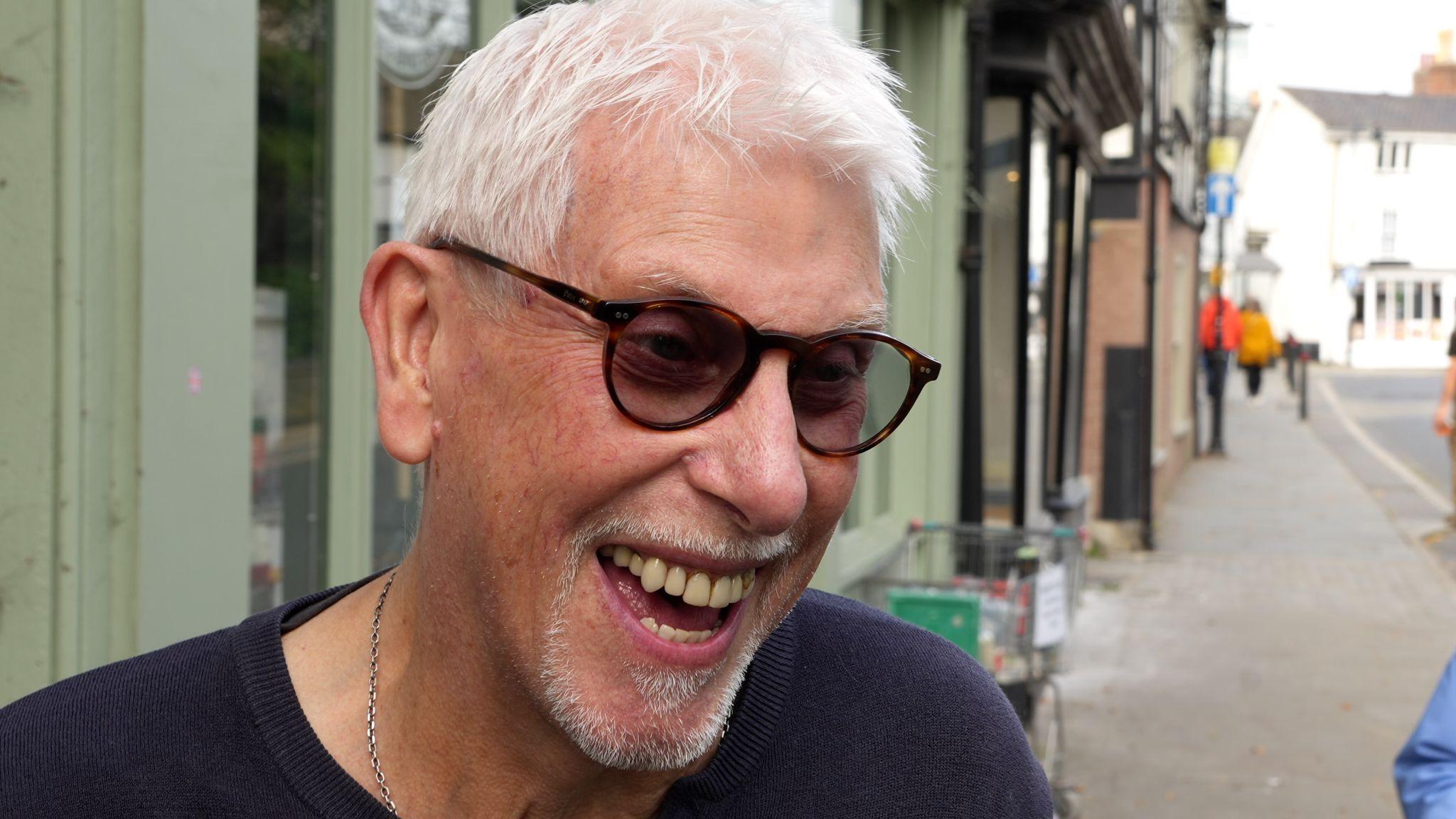 Roy is smiling and has short, white hair. He is wearing sunglasses and a dark top. There are shops in the background