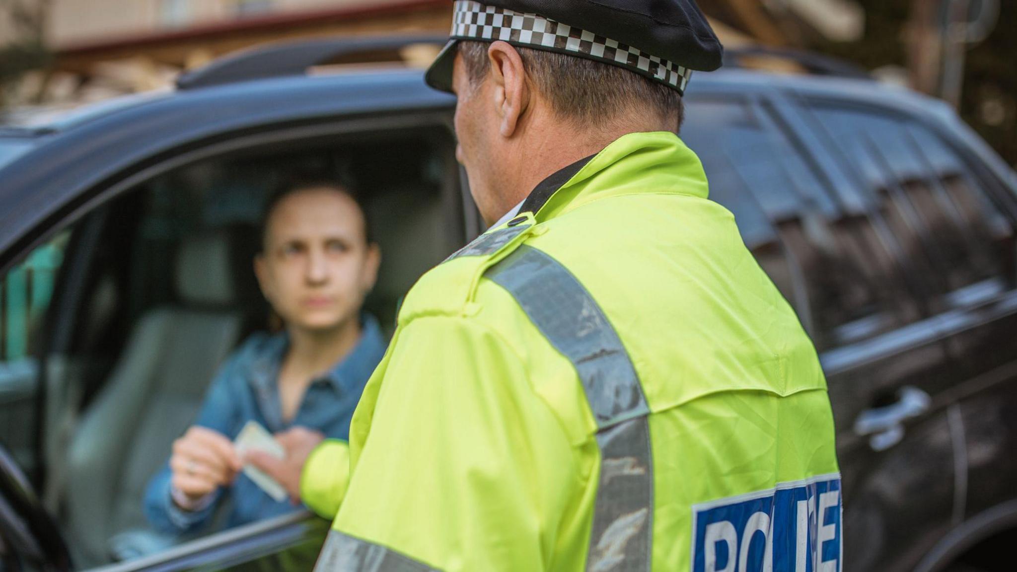 A policeman wearing a yellow hi-vis jacket and a black and white chequered hat. He is speaking to a woman driving a black car, and passing her license back to her through the open window.