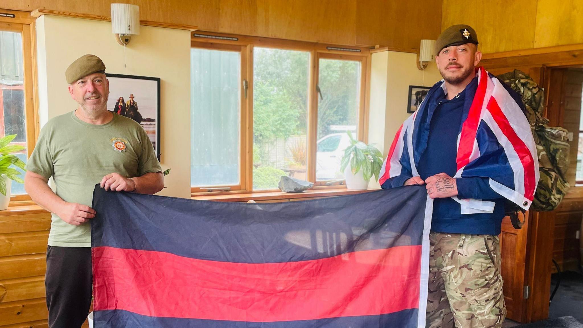 Jonathan Farrelly and another man holding up a flag. Jonathan also has a Union Flag around his shoulders and is wearing a military uniform.