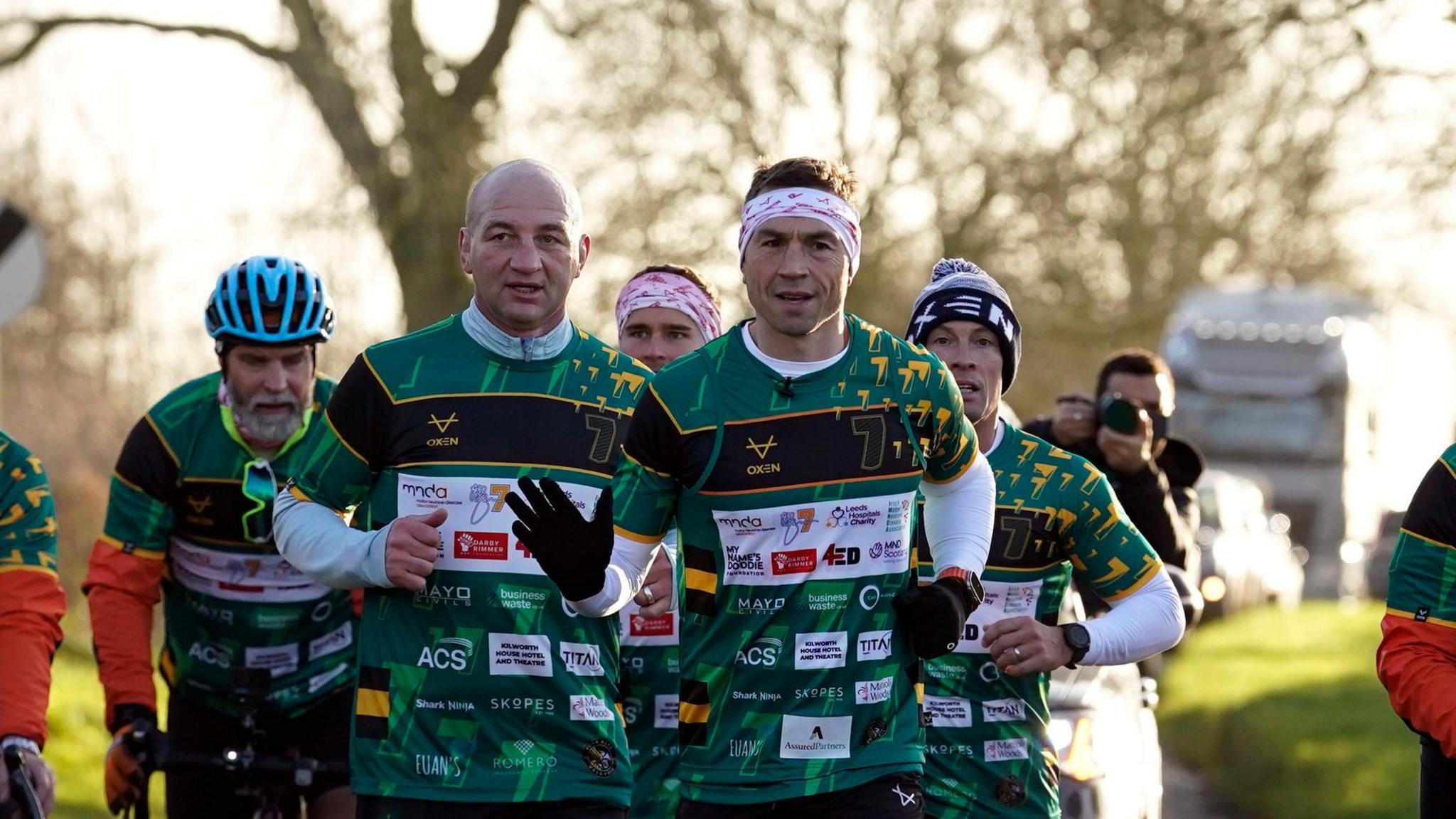 A pack of runners all wearing green and black rugby shirts run down a road with a car behind them. Three people on bicycles cycle alongside them. 