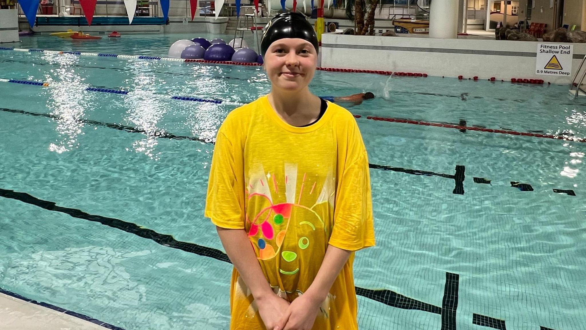 A girl in a large, yellow Pudsey shirt and a black swimming cap on. There is a pool behind her