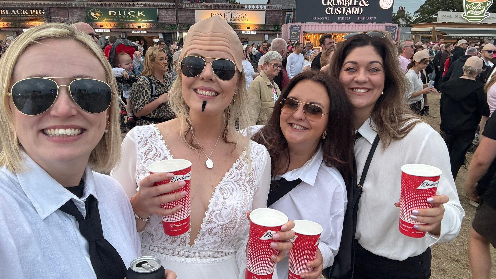 Four women enjoying a music festival. Kym is in the middle, wearing a white wedding dress, bald cap and stick-on goatee. Her friends have white shirts and black ties, and two have sunglasses. They are holding red drinks cups.