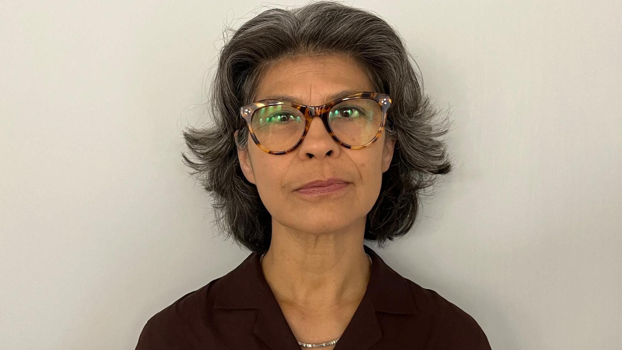 Close-up portrait of Sarah Armstrong wearing round tortoiseshell glasses and a dark collared top, facing the camera against a plain light-colored background.