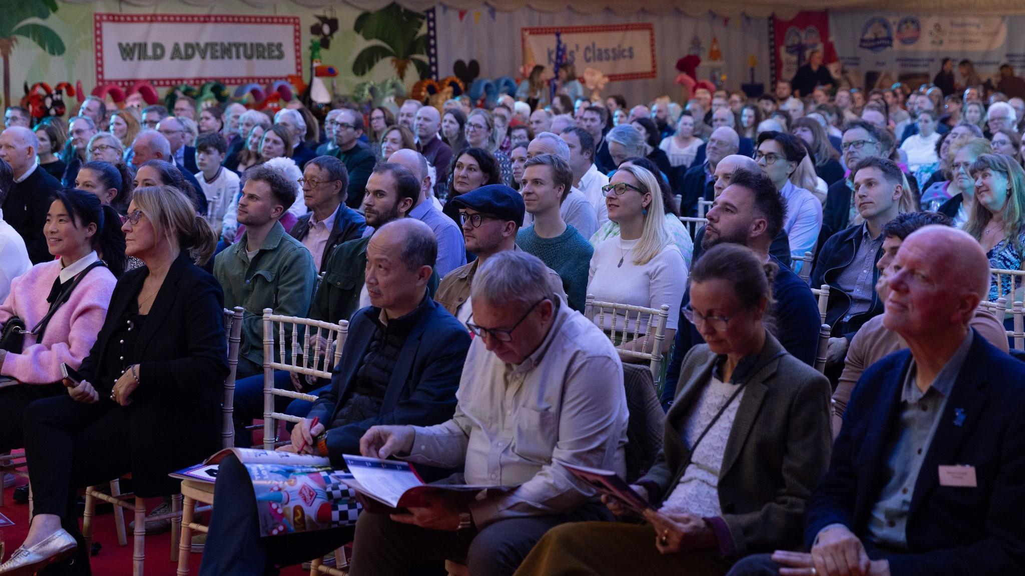 A dimly lit events space with around a hundred people sitting in chairs, looking up at the auctioneer on stage at the front of the room. 