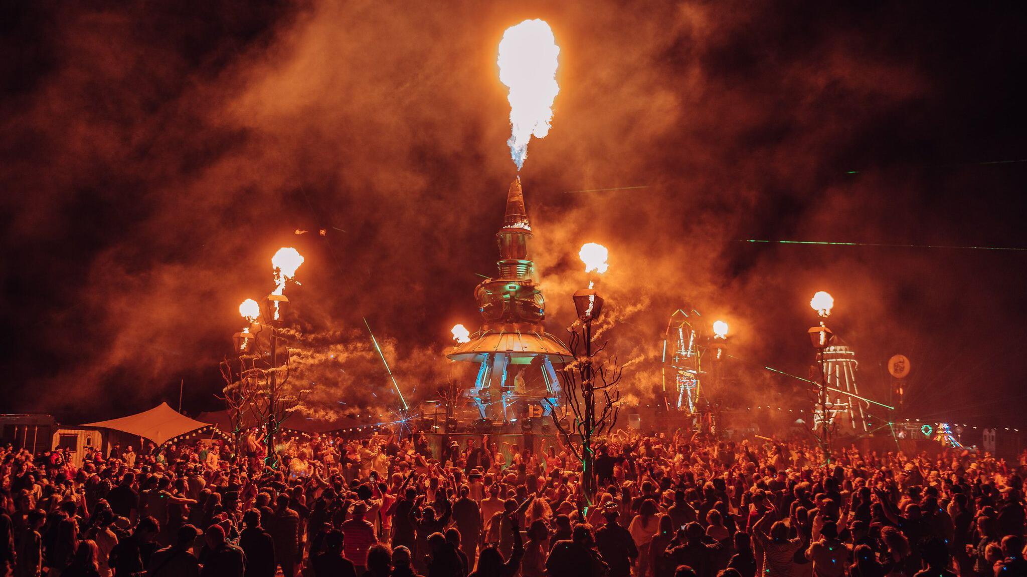 A crowd standing around a giant metal installation shooting flames