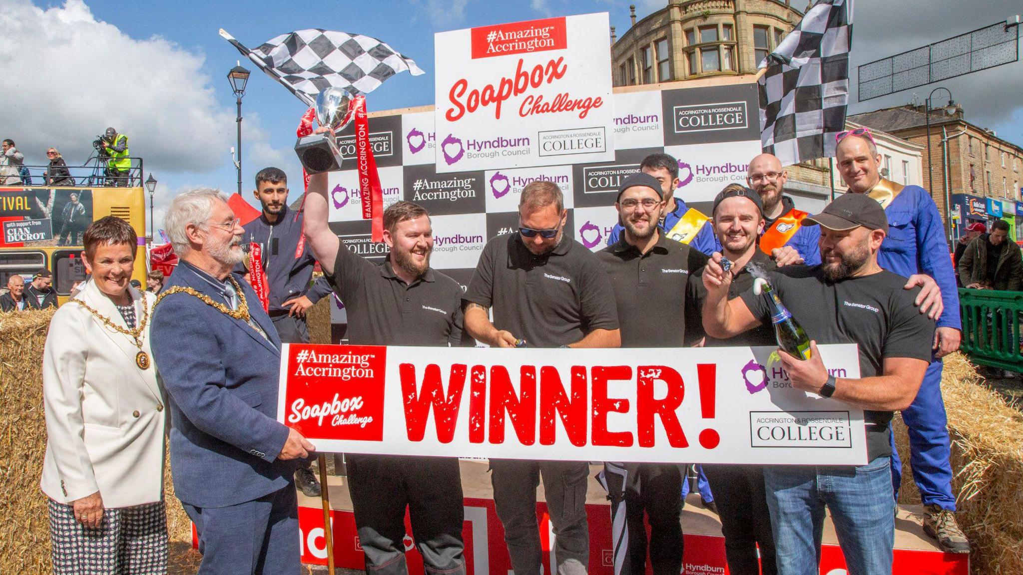 Members of the winning team smile and hold a trophy, as well as a banner saying 'Amazing Accrington Soapbox Challenge winner!' They are standing next to the Mayor and Mayoress of Hyndburn.