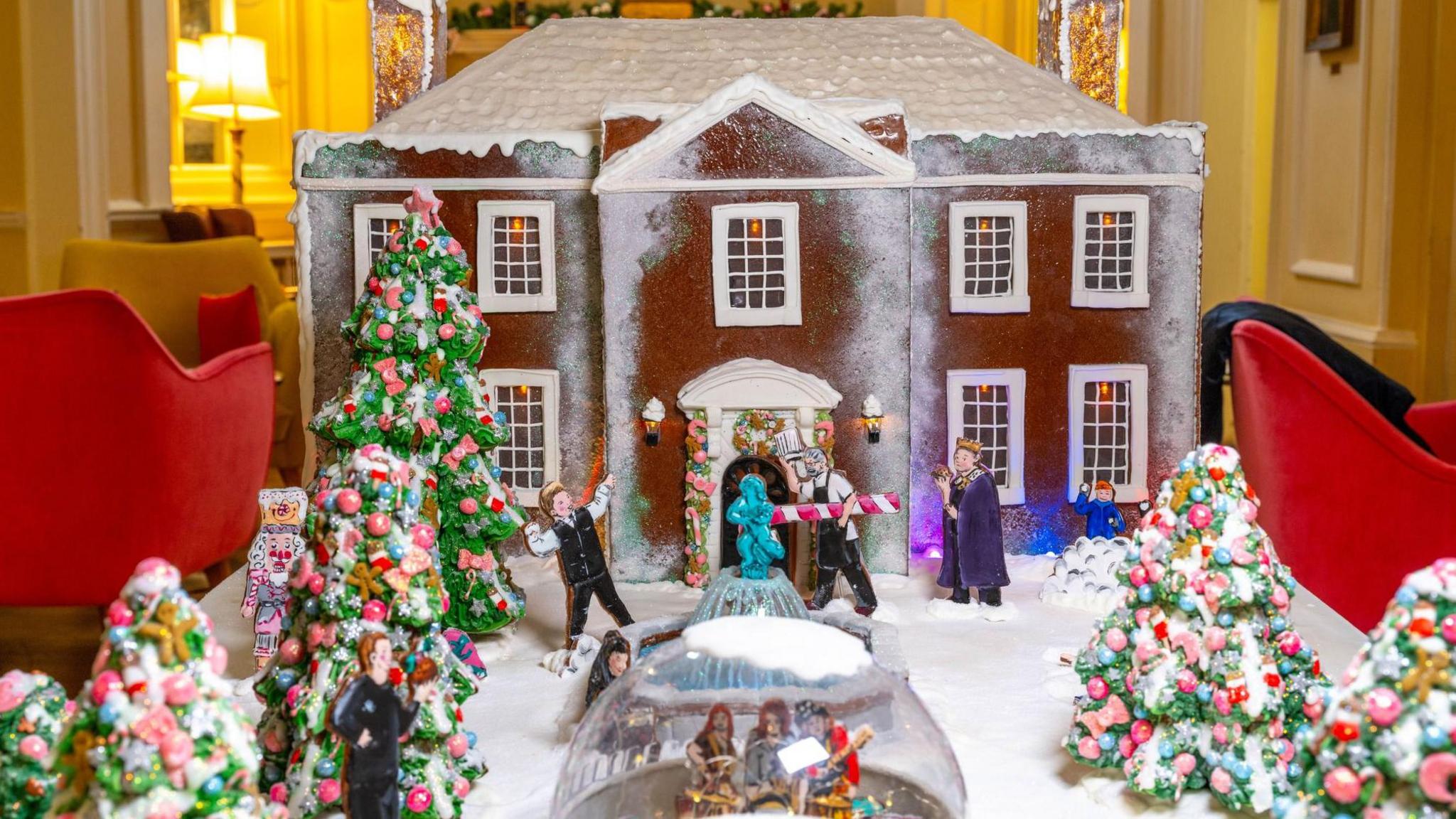 A close-up shot of the gingerbread model, which shows a Georgian-style house topped with snow and decorated Christmas trees and two igloos in its snowy front garden, laid out on a table inside The Assembly House and surrounded by chairs.