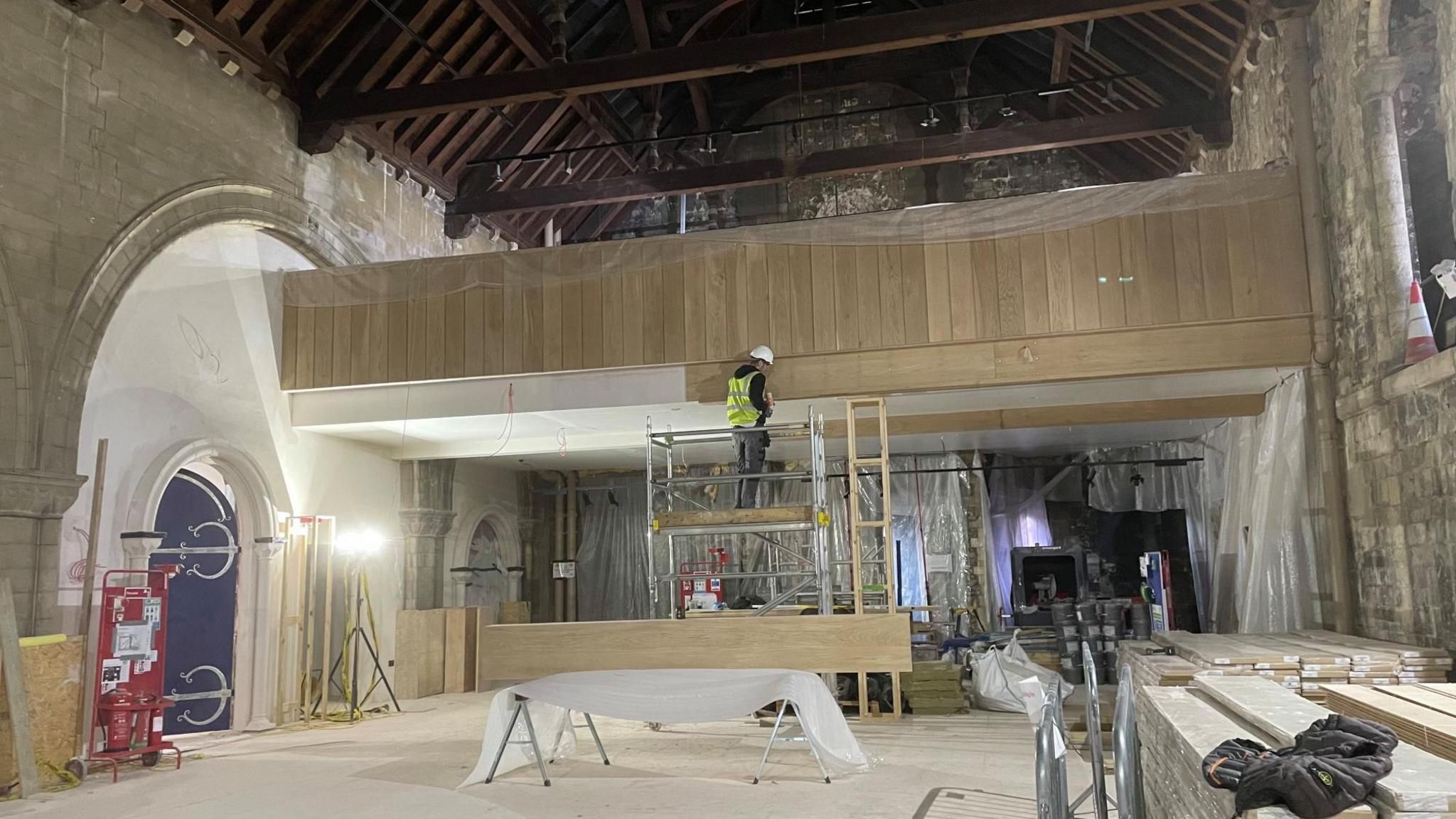 Work being completed to the interior of Norwich Castle. A builder stands on scaffolding and appears to be fixing wood panelling to a wall. Stacks of wood are piled up on side of the room