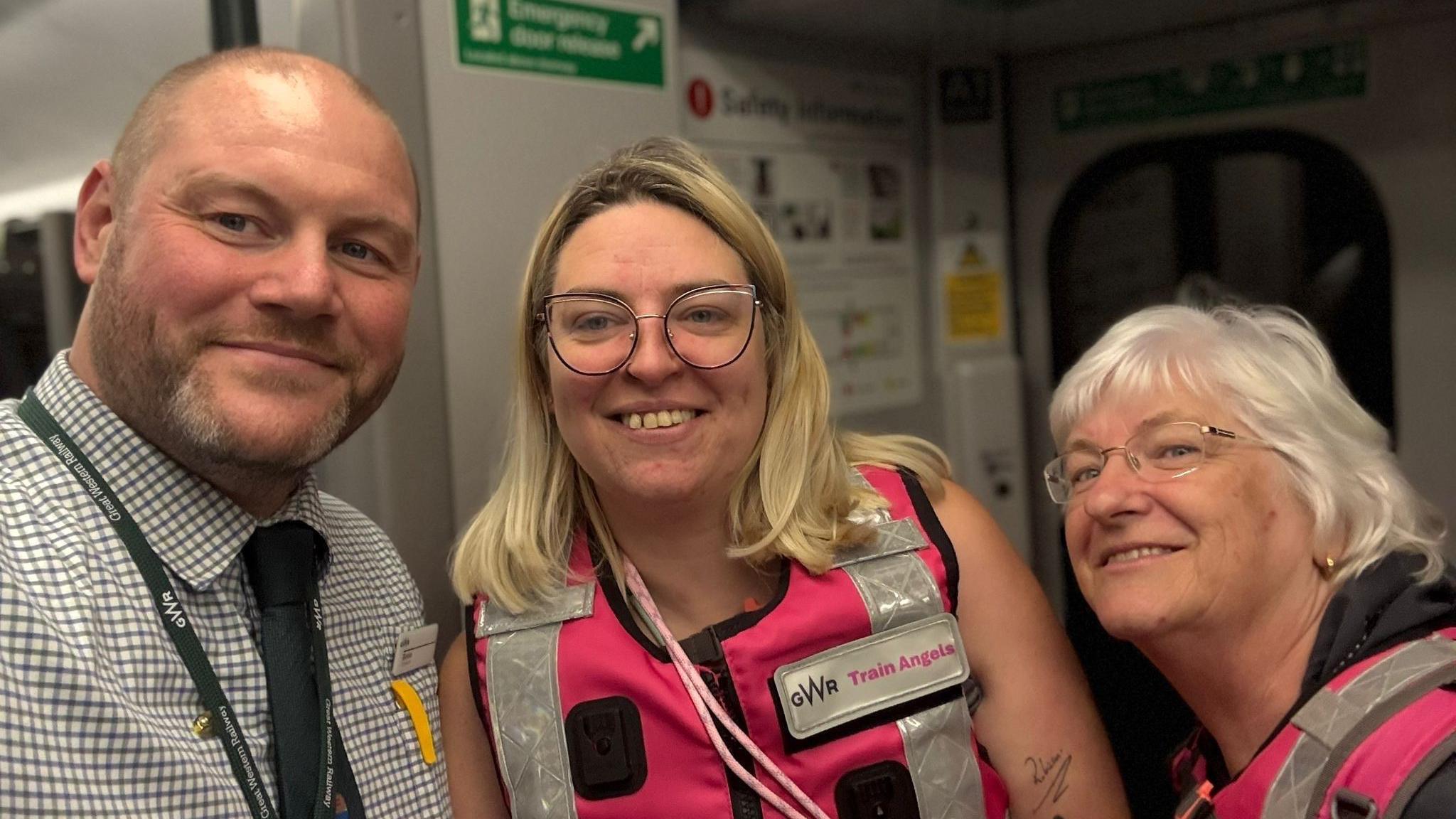 Two Train Angels in pink high-vis and glasses stand alongside a GWR employee inside a train carriage, all are smiling at the camera.