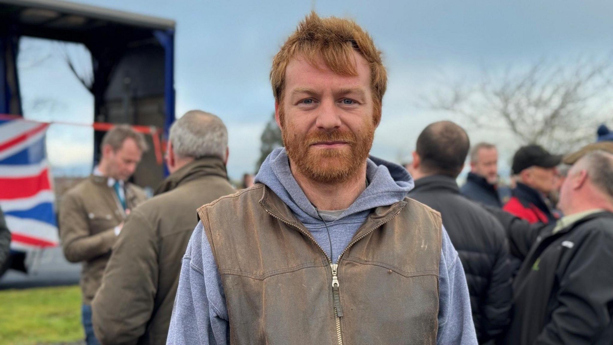 Ben Leyland is a man in his 30s with ginger hair and beard. He is wearing a blue top and there are other farmers standing behind him 