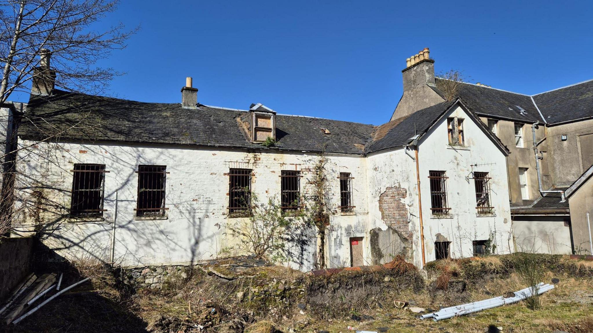 A large derelict building with crumbling white washed render. There are boarded up windows and the area next to the building looks overgrown and abandoned.