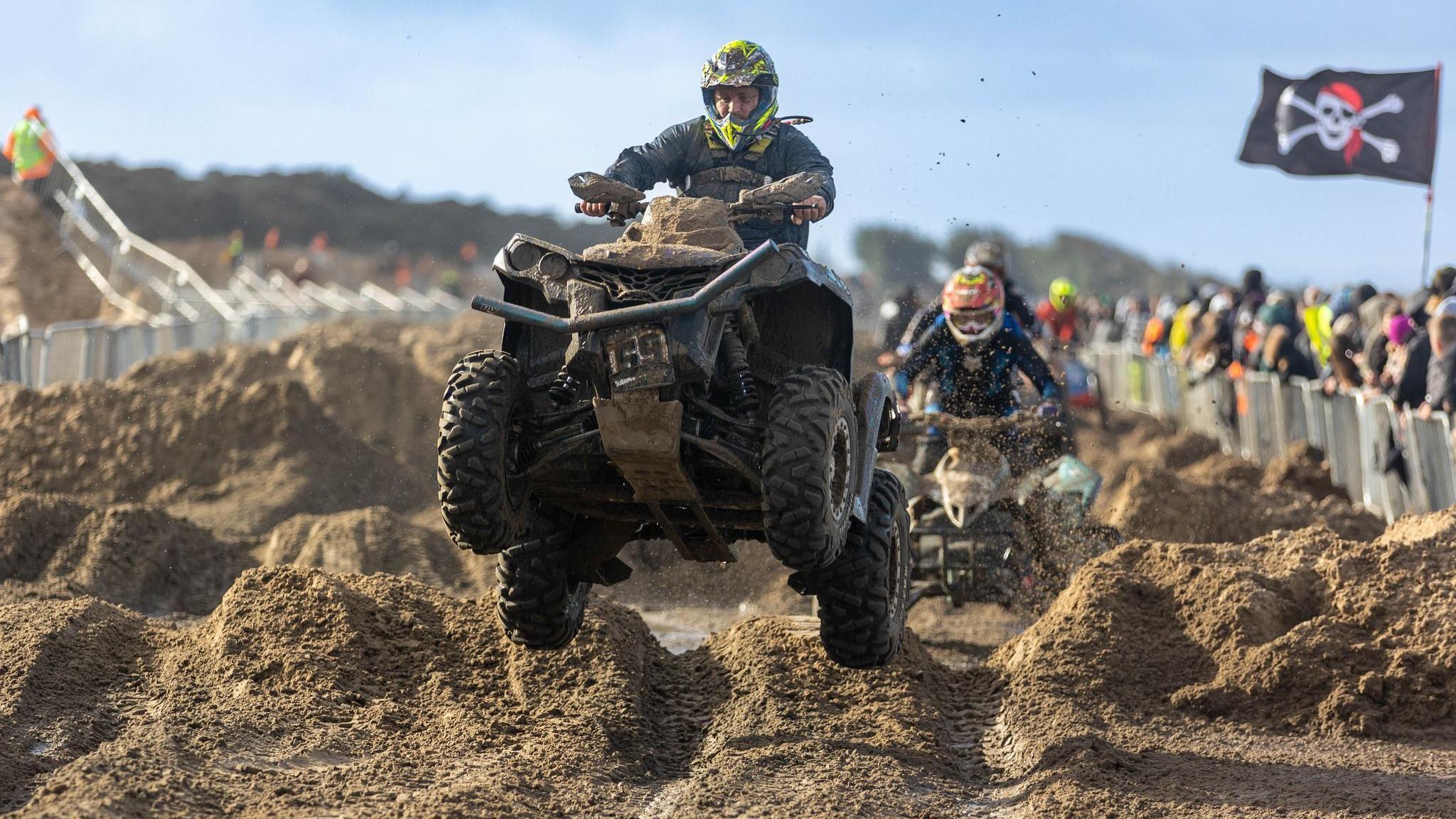 A man on a quad bike goes over the sandy hills of the Weston Beach Race course. He is wearing professional racing leathers and a crash helmet. Other contestants are visible behind him, along with spectators, one of them has a large pirate skull and crossbones flag on a pole