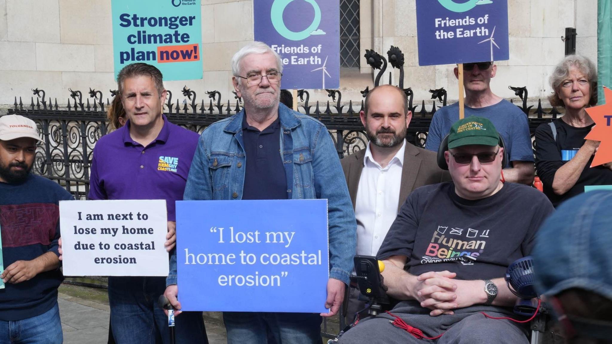 A group of people are standing outside the High Court in London. One of them, Kevin Jordan, has a sign saying, "I lost my home to coastal erosion", and another person to his right has a sign saying "I am next to lose my home due to coastal erosion".