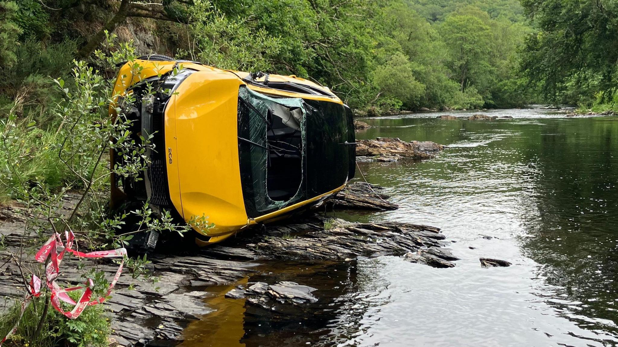 The yellow car is lying on its side next to the River Dart. The windscreen is cut away and smashed, the bumper is ripped away and the front and top are bent. There is red and white incident tape wrapped around a nearby bush.