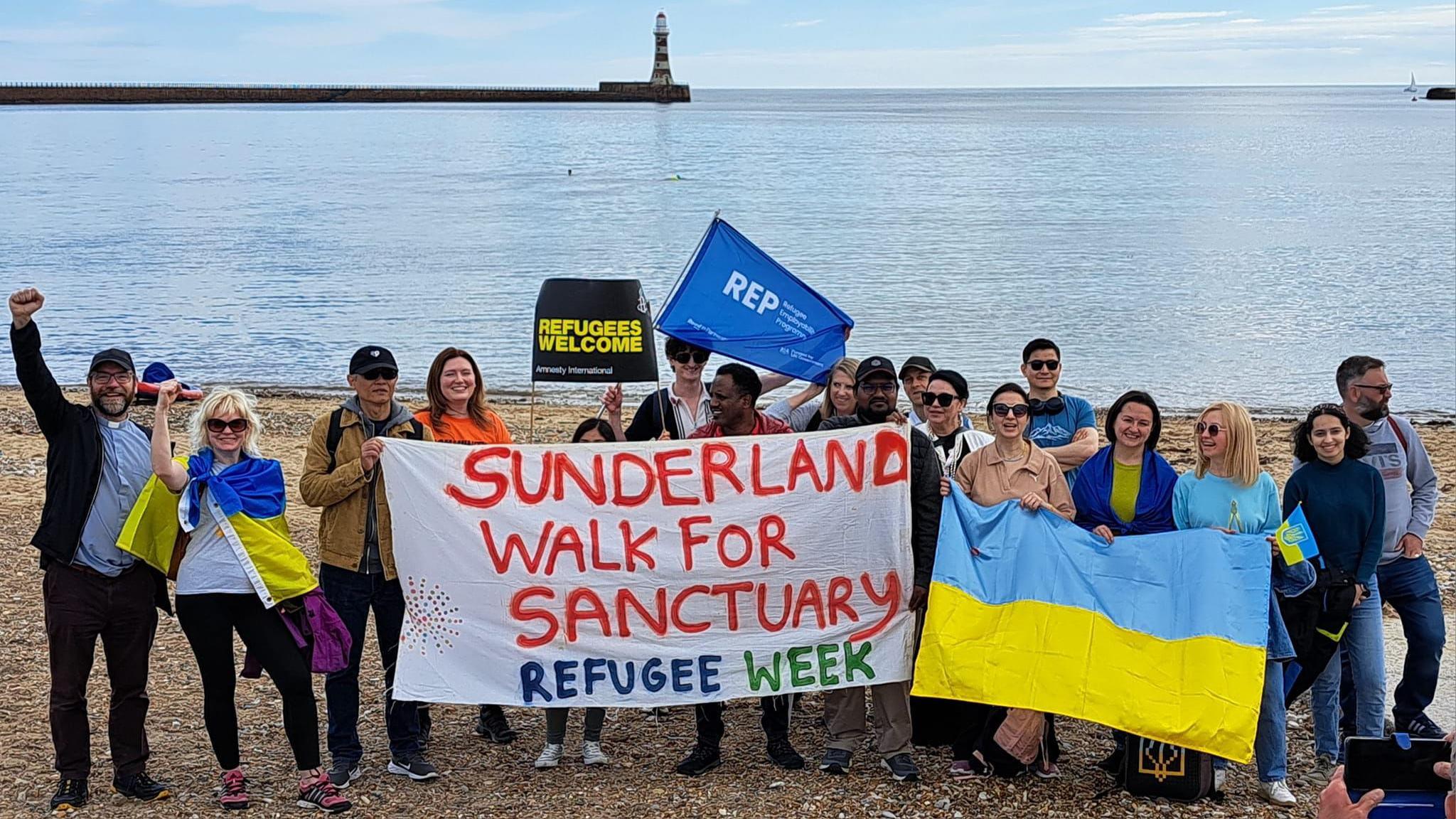 About 20 people holding Refugees Welcome sign, an Ukrainian flag and a Sunderland Walk for Sanctuary Refugee Week flag at Roker Beach in Sunderland. Roker Pier and lighthouse are visible in the background and there is a single boat in the calm blue sea.
