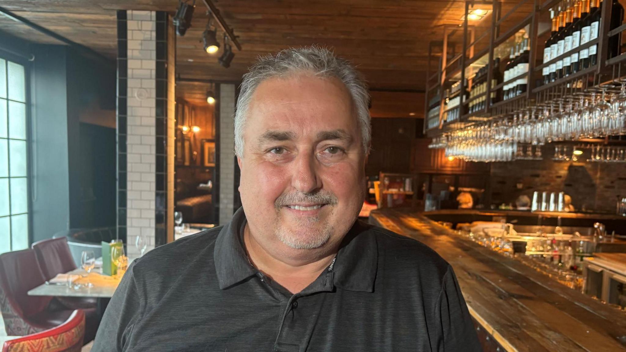 A man with short grey hair and a short grey goatee beard looks into the camera. He wears a grey polo shirt. Behind him is a wooden bar, over his left shoulder and white tables and maroon chairs over his left shoulder.
