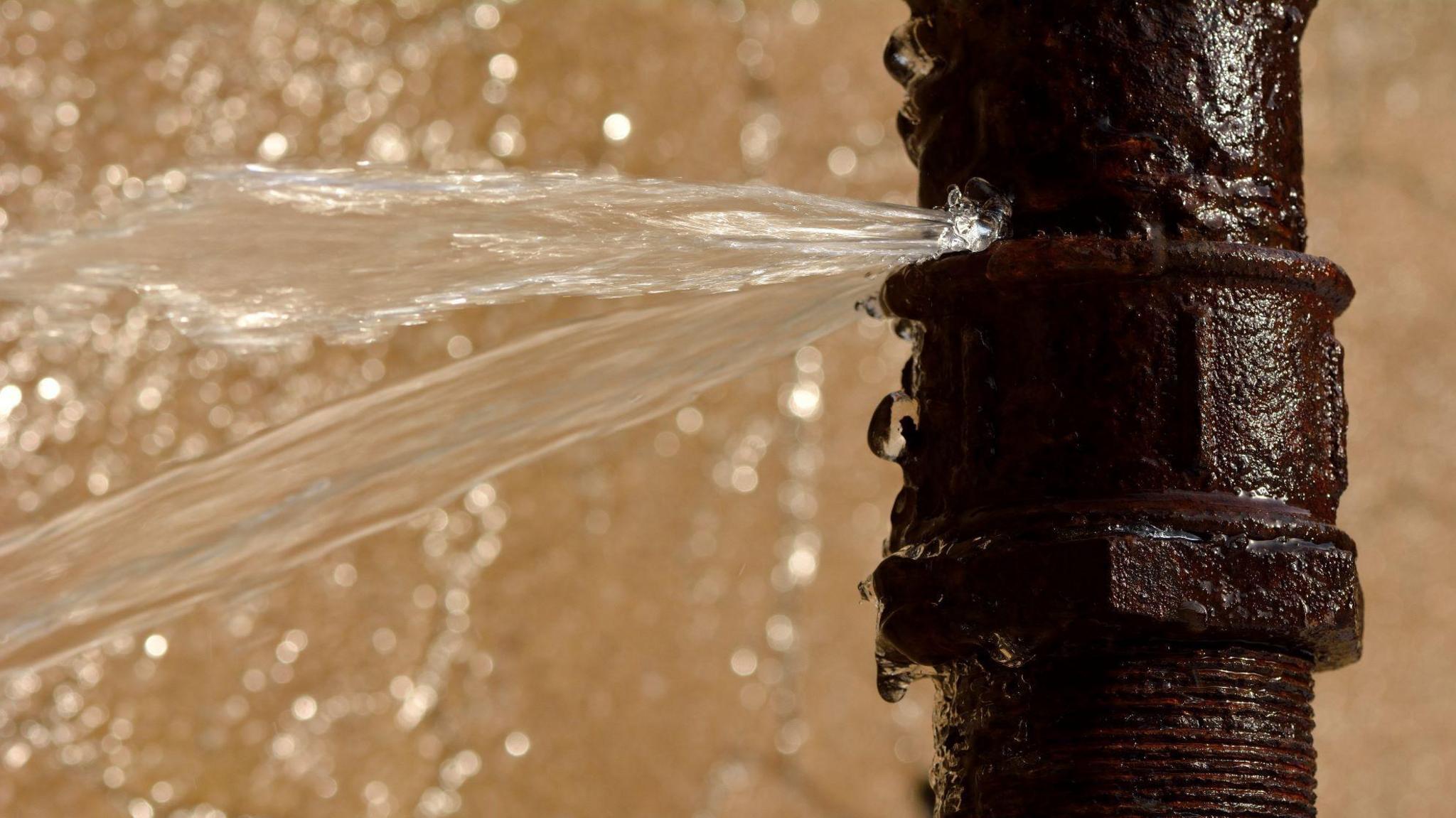 A stock image of a burst water pipe. The pipe is black and water is spraying out of it. 