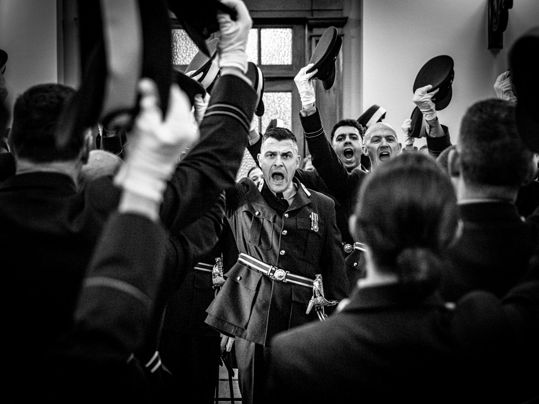 A black and white image of officers holding their caps in the air as a man centre stage walks down the isle.  The three officers facing the camera are either cheering or shouting and the photo captures the moment.